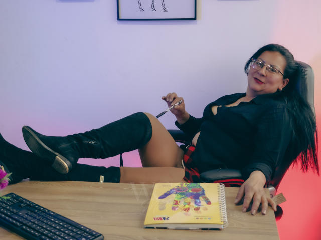 MissLadyy with glasses, black top, red plaid skirt, black boots, and bare thighs, sitting on desk, holding pen, smiling, notebook with colorful doodles, keyboard in foreground.