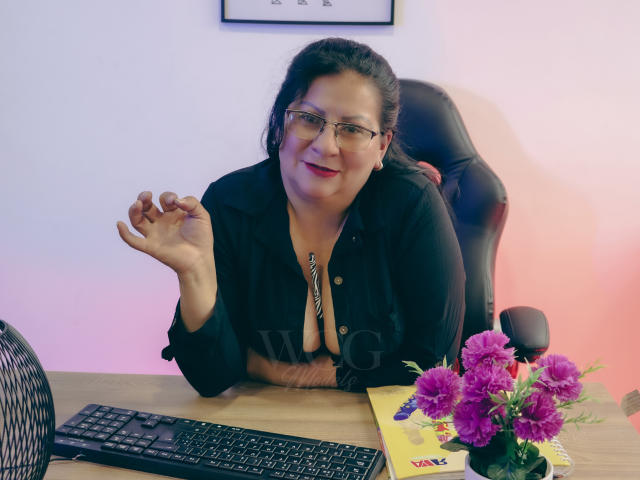 MissLadyy, chubby, dark-haired, glasses, black shirt, cleavage, hand up, smiling, desk, keyboard, flowers, office chair, light purple-pink wall, red lipstick, serious yet friendly.