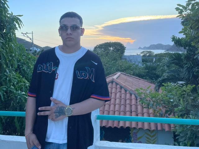 ZairaAndres stands in front of a rooftop, wearing a black baseball jersey with "LA" in white letters and red stripes, over a white t-shirt. He has short dark hair, glasses, and a tattoo on his right hand. He wears a gold watch on his left wrist. The background includes greenery, a tiled roof, and a distant ocean with mountains. The sky is blue with an orange sunset. The image is clear and focused.