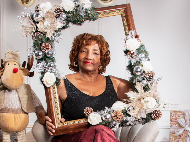PatriciaLaens with curly red hair, wearing a black sparkly top, holding a large Christmas wreath frame with pine, silver, and white decorations, sitting next to a reindeer stuffed toy.