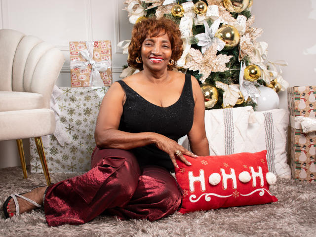 PatriciaLaens with curly brown hair, medium brown skin, black sparkly top, red satin pants, sitting on carpet, smiling, holding red "HoHoHo" pillow, Christmas tree and gifts in background.