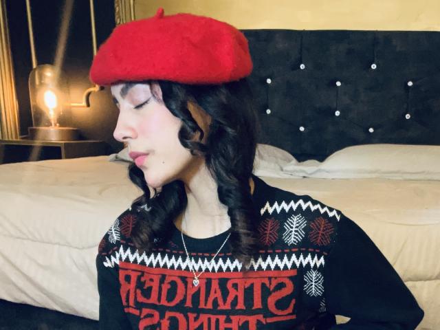 AitanaJohns has curly brown hair, a red beret, and a black sweater with red and white patterns. She's smoking a cigarette, with a bed in the background.