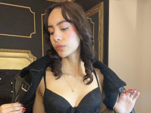 AitanaJohns with curly brown hair, wearing a black lace bra, zipping up a black jacket, showing her red-painted nails, and a delicate necklace with a small heart.
