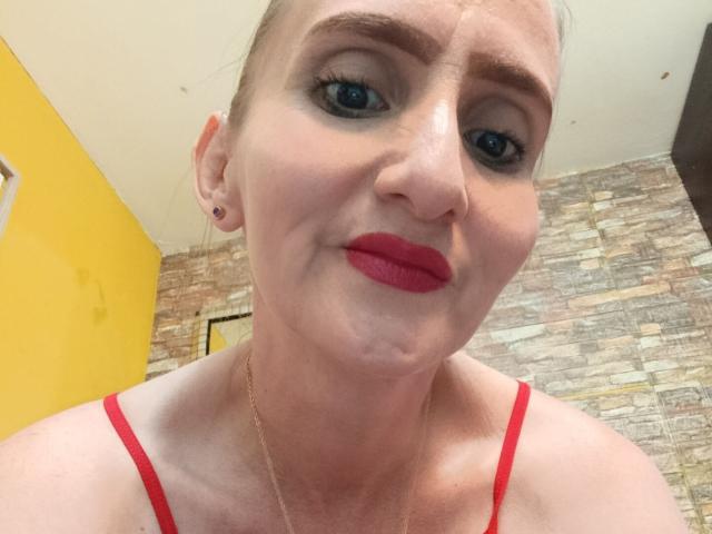 RubiAnderson has fair skin, blue eyes, red lipstick, and red bra straps, with a gold necklace and small black earrings, looking up with a slight smile.