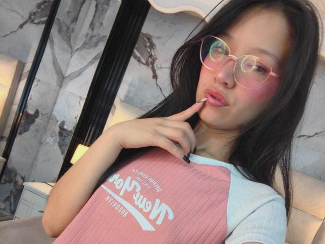 SophieDelaney with pink glasses, pouty lips, finger on lip, wearing pink and white raglan shirt, black hair, marble wall, black nail polish, looking cute.