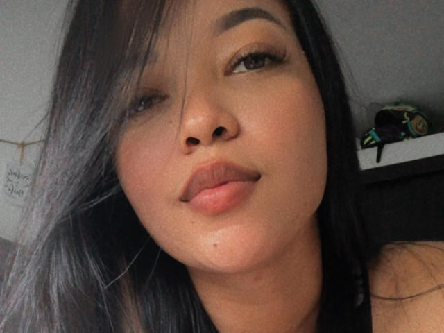 BellaGrin's face, with long black hair, slightly parted lips, and a small mole below her chin. She has brown eyes, dark eyebrows, and a soft smile.