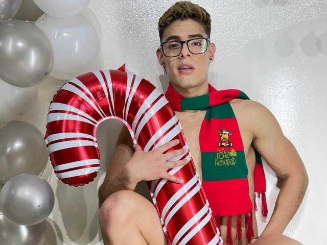 AngelEvil, wearing glasses and a green-red striped scarf, holds a shiny red and white candy cane balloon, sitting with a muscular, shirtless upper body, short blonde hair.