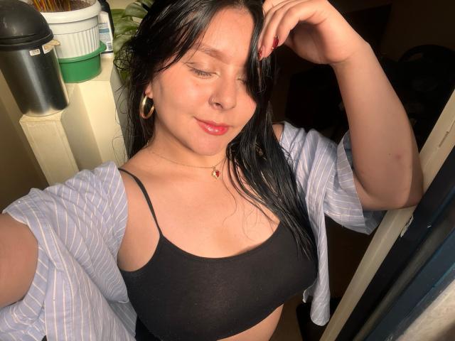 EmaJames with wet black hair, wearing a black sports bra, blue striped shirt, gold hoop earrings, red lipstick, and a necklace, smiling at the camera, showing her tits.