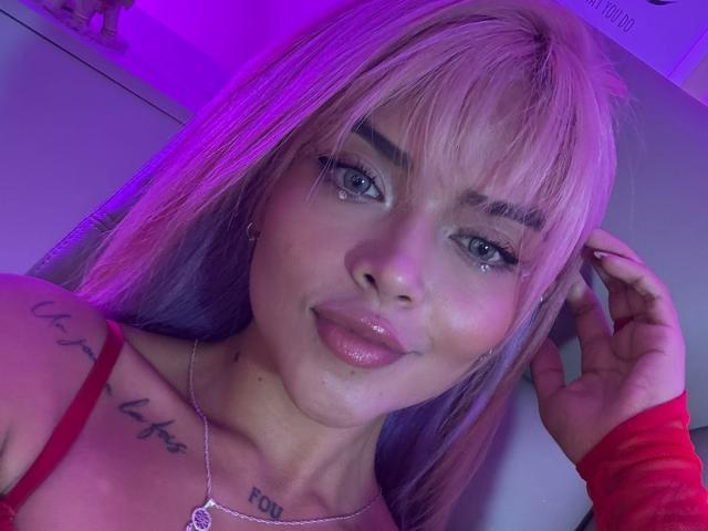 MajoJones with pinkish-purple hair, blue eyes, glossy pink lips, red top, visible tattoo on right shoulder, holding red sleeve, purple and pink lighting.