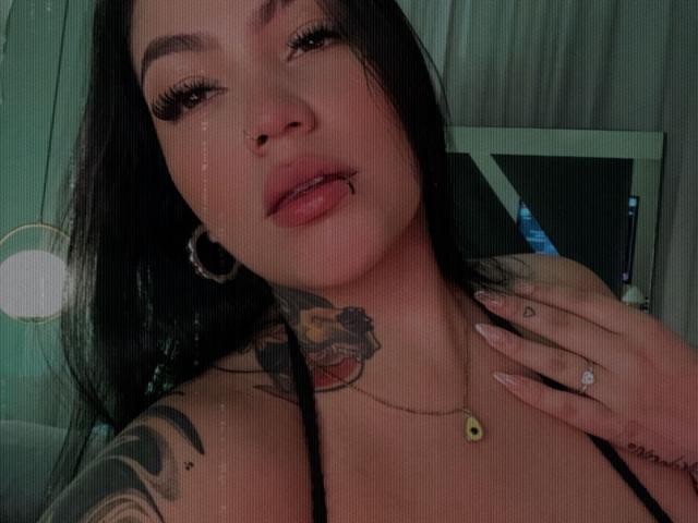 TessaMagic has long black hair, a nose ring, and a lip piercing, with a tattoo on her neck, wearing a black top, and showing her hand with a ring.