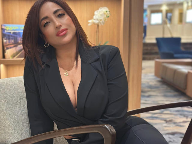 AbbySprouse with medium brown hair, wearing a black blazer with deep cleavage, showing her big boobs, sitting in a chair, looking slightly to the side, smiling.