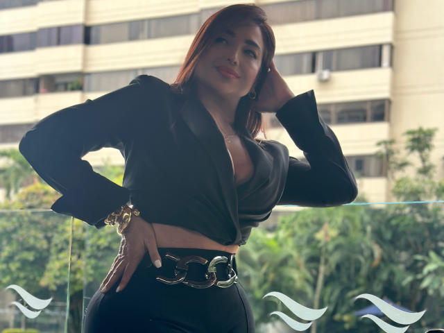 AbbySprouse with red hair, black blazer, deep cleavage, black pants, gold belt, hand on hip, glass balcony, wrist bracelet, confident smile, city building background, daytime, sexy.