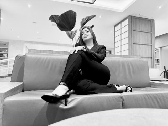 Black-and-white photo of AbbySprouse with long dark hair, sitting on a leather couch, holding large poppy flowers, wearing a black suit and heels, legs crossed, looking up.