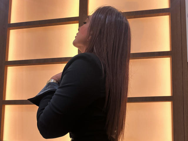 AbbySprouse with long brown hair, black top, arms crossed, side profile, looking up, golden light behind, wearing a silver watch, elegant and calm.