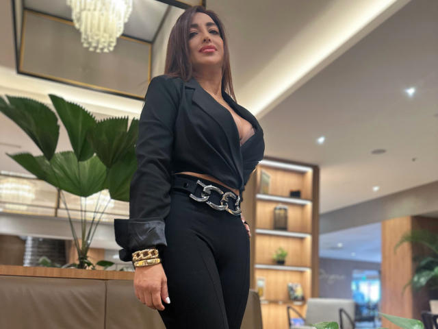 AbbySprouse with big boobs, wearing a black blazer, exposing her tits, black pants, and a belt with a silver buckle, standing in a modern room with a chandelier.
