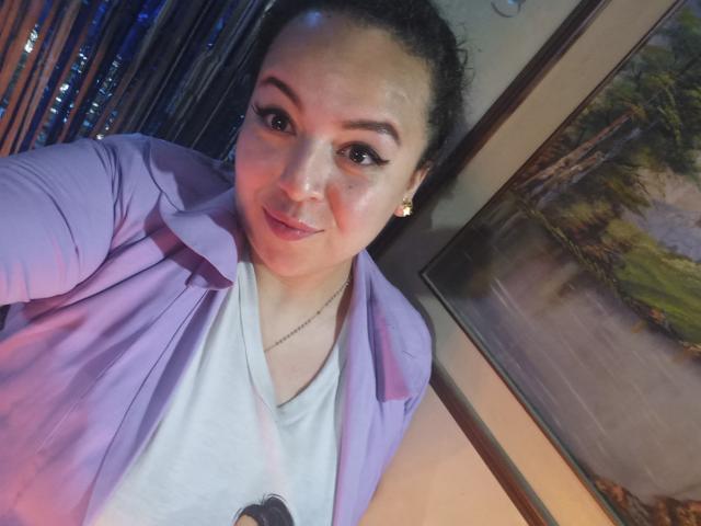 NatashaAmbeer, with curly dark hair, pink jacket, white tee, and subtle makeup, has a cute smile and small earrings, showing her face in a selfie.