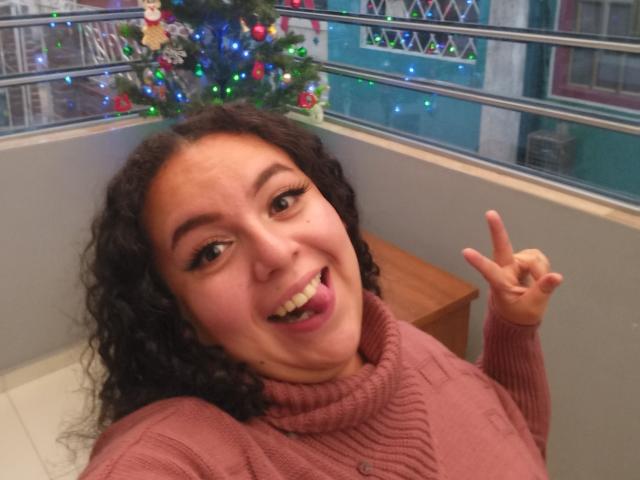 NatashaAmbeer with curly brown hair, pink turtleneck, and a playful expression, sticking out her tongue and making a peace sign with her right hand in a selfie.