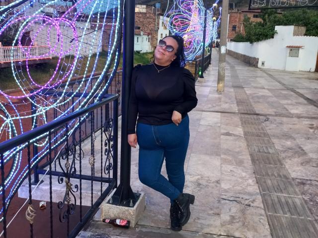 NatashaAmbeer with big ass, wearing a sheer black top showing her tits, blue jeans, and black boots, standing by colorful neon lights, wearing glasses, and looking up.