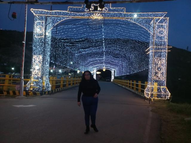 NatashaAmbeer stands in front of a glowing, blue-lit archway with white lights, wearing a black top and blue pants, with her ass slightly turned to the camera.