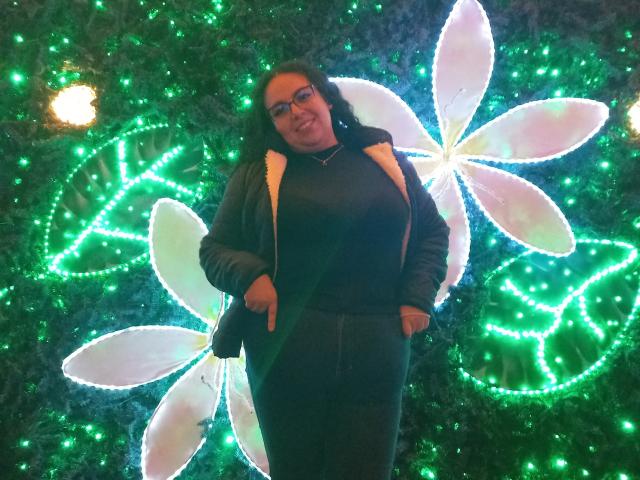 NatashaAmbeer with glasses, black hoodie, and dark pants, standing in front of a glowing green and white floral light installation, hands in pockets, smiling.