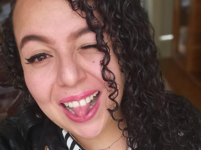 NatashaAmbeer's smiling face with one eye closed, curly black hair, pink lipstick, white teeth, black eyeliner, and a black leather jacket. Her tongue is slightly out.