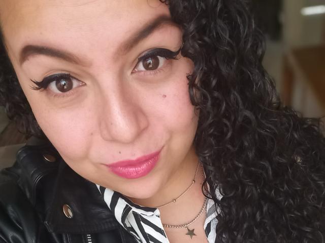 NatashaAmbeer with curly black hair, brown eyes, and pink lipstick, wearing black eyeliner and a black leather jacket, smiling at the camera. She's also wearing a zebra-patterned shirt.