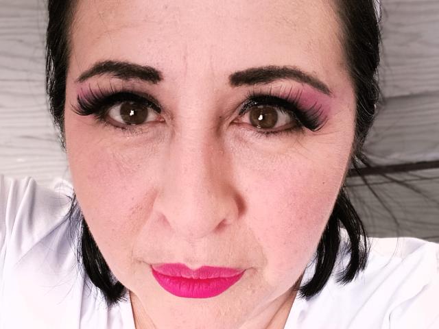 Close-up of ScarletMilfX with light skin and dark brown eyes. She has thick black eyebrows, long black eyelashes, and pink eyeshadow. Her lips are painted bright pink. She wears a white shirt, and her black hair is slightly messy. The image focuses on her face, showing fine skin texture and slight freckles. The composition is centered, with her eyes looking directly at the camera. The overall style is realistic and detailed.