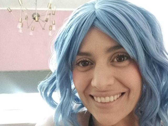 LilaaSalmon has blue wavy hair, brown eyes, and a smiling face with white teeth. She's wearing a black top, and a modern chandelier is in the background.