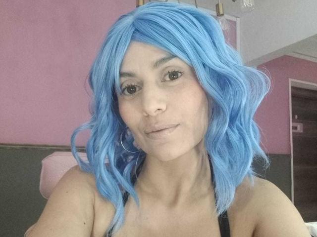 LilaaSalmon with blue wig, pouty lips, and hoop earrings, showing her tits in a black tank top, looking at the camera. Her ass is not visible.