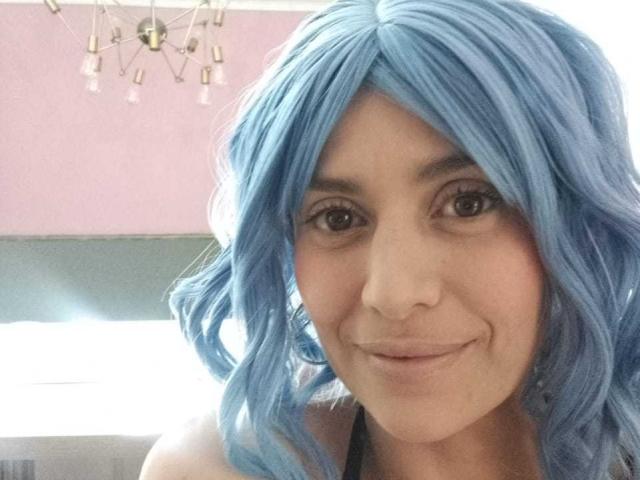 LilaaSalmon with blue hair, smiling, showing her face, small nose, and brown eyes. She's wearing a black top, and has wavy blue hair. Her tits are not visible.