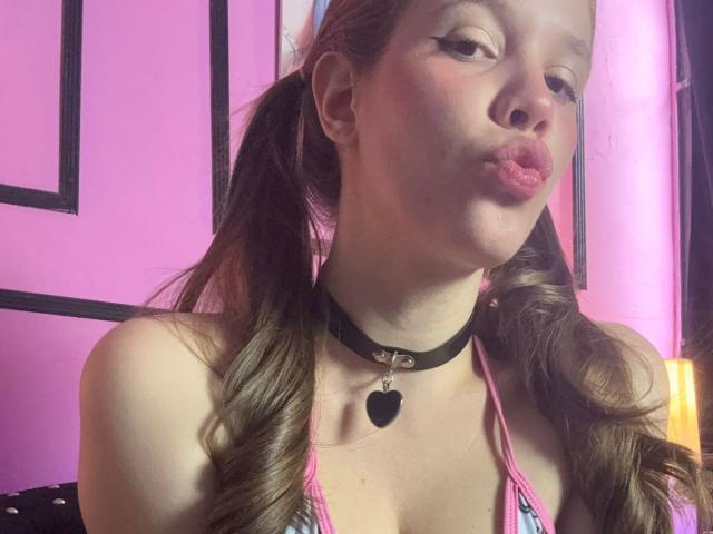 ArielLoveX with pigtails, pink bikini top, black choker with heart, puckering lips, pink walls, and a yellow lamp in the background. She's a sexy, cute, and attractive woman.