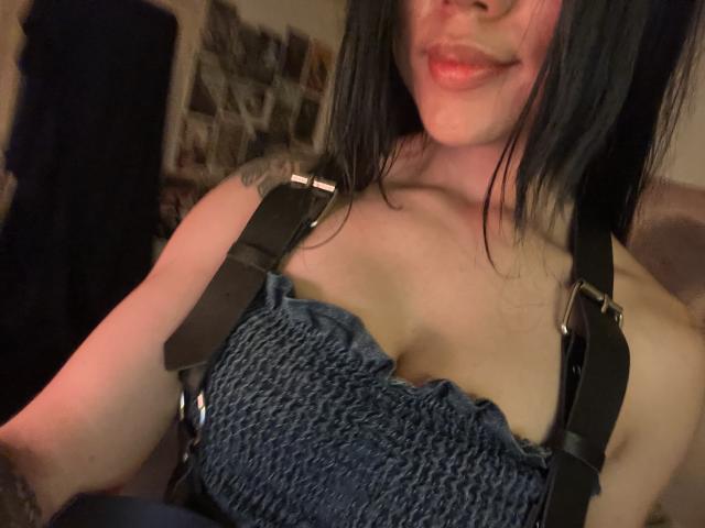 SteffanyTaylor with black hair, wearing a blue, textured, strapless top with black straps, showing her tits and a tattoo on her left arm, smiling with red lips.