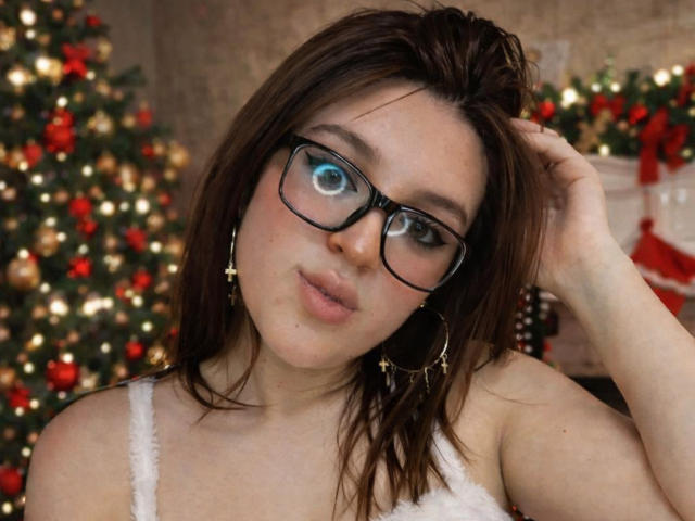ZoeyClark with glasses, brown hair, and hoop earrings, slightly smiling, hand in hair, wearing a white fuzzy top, blue eyes looking to the side. Boobs visible, Christmas decor background.