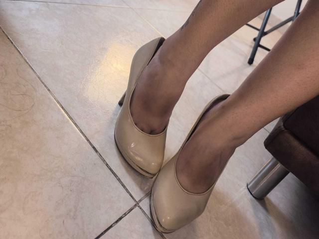CarolineVoss's legs in shiny beige high heels with a slight heel, standing on tiled floor, her skin is light, and a metal chair leg is partially visible.