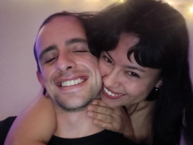 TantricLovers with short dark hair and slight beard, smiling with eyes closed, has arm around TantricLovers with long black hair, smiling, both showing teeth. TantricLovers' hand rests on TantricLovers' shoulder.