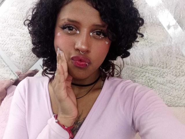LunaKeller has curly black hair, a nose ring, and pink lipstick. She's pouting with her finger on her cheek, wearing a pink top and a black choker.