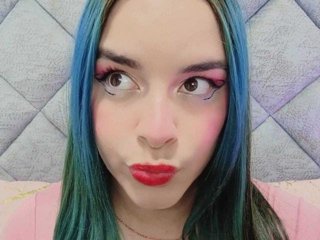 AuroraSantorini with blue-green hair, pink eye makeup, and red lipstick, puckering her lips in a selfie. Her brown eyes look slightly to the side.