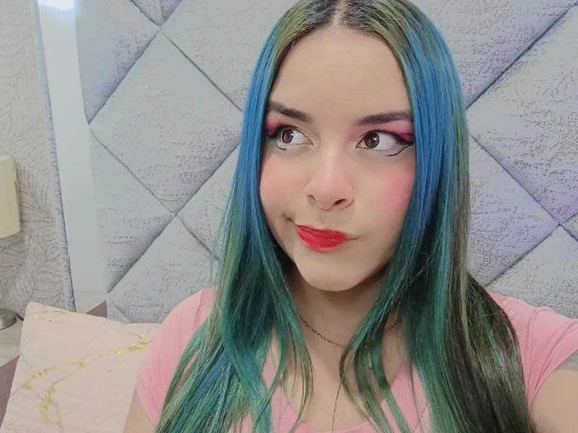 AuroraSantorini has blue-green hair, pink eyeshadow, red lipstick, and a cute smile. She's wearing a pink shirt and looking to the side with a playful expression.