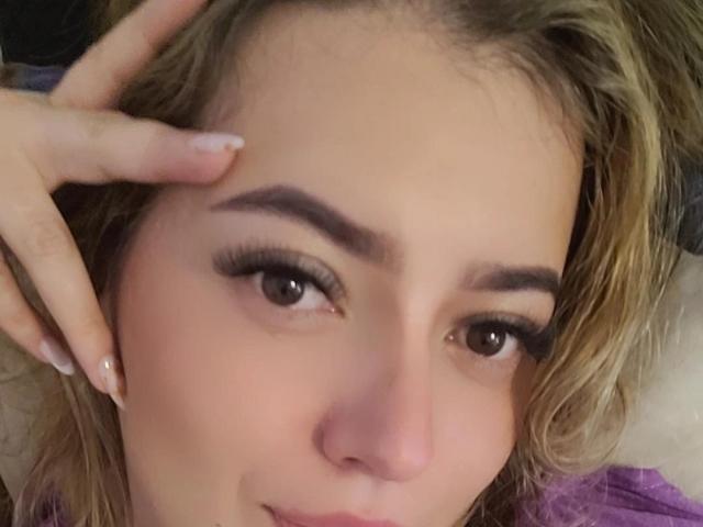 Close-up of KristenSexy's face with light skin, blonde wavy hair, and brown eyes. She has thick, dark eyebrows and long, black eyelashes. Her right hand, with neatly manicured nails, rests on her forehead. She's looking directly at the camera with a slight, confident smile. The background is blurry, with a hint of purple and beige. The image has a soft, natural light that highlights her facial features. The texture of her hair is slightly wavy and loose. KristenSexy's facial expression is relaxed and slightly playful.