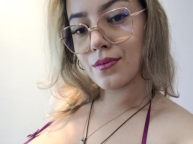 Close-up of KristenSexy, a light-skinned woman with blonde, wavy hair. She wears large, square, gold-rimmed glasses and has purple lipstick. Her eyebrows are dark and neatly shaped. She has a slight smile and is looking directly at the camera. She wears a thin, black cord necklace with a small pendant and a maroon halter top. The image is well-lit with soft, natural light highlighting her face.