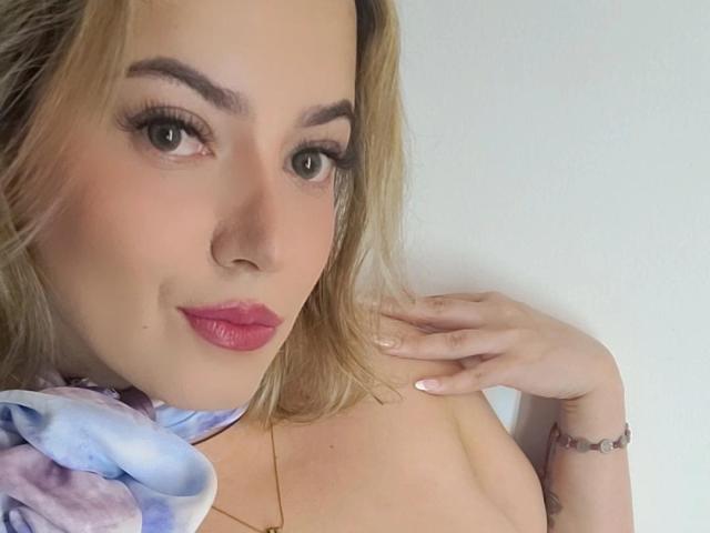 KristenSexy has blonde hair, green eyes, and pink lipstick. She's wearing a colorful scarf, has a bracelet, and is touching her neck with a hand. Boobs visible.