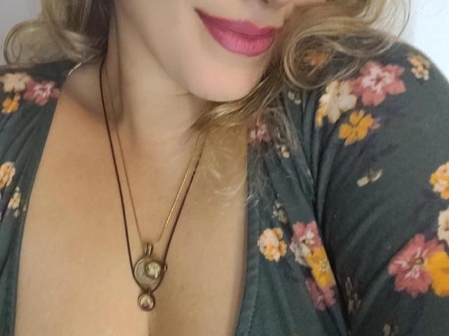 KristenSexy's face with pink lipstick, blonde curls, wearing a floral green top, necklace with heart-shaped pendants, showing cleavage, soft skin, partial view of her tits.