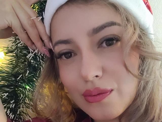 Close-up of KristenSexy with wavy blonde hair, wearing a red Santa hat. She has dark eyebrows, brown eyes, and pink lipstick. Her right hand, with long, glittery nails, touches the hat. A Christmas tree with green needles and yellow lights is in the background. Her skin is fair, and she has a small nose ring. The image focuses on her face, showing a slight, confident smile. The composition highlights her facial features and the festive hat.