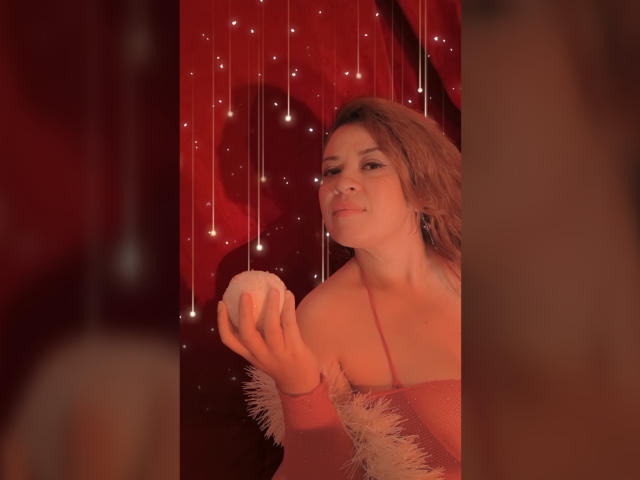 RosseHot69 con i capelli rosa, che tiene una palla bianca, indossa una maglietta rosa con maniche vaporose, appare carina e sicura di sé, su uno sfondo rosso, scintillante..