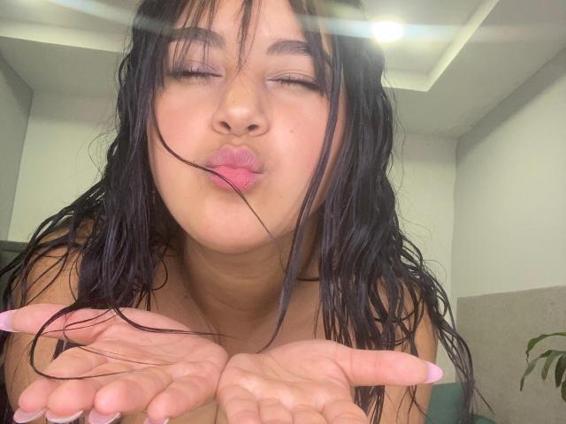 Auroratziu with wet black hair, eyes closed, pouting pink lips, hands up, in a selfie. Sunlight flares in the top. Auroratziu's tits are slightly visible.