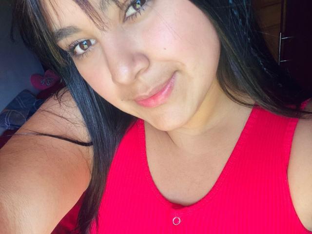 Auroratziu with light brown skin, black hair, green eyes, and a red ribbed tank top, smiling slightly at the camera, showing a subtle nose and pink lips.