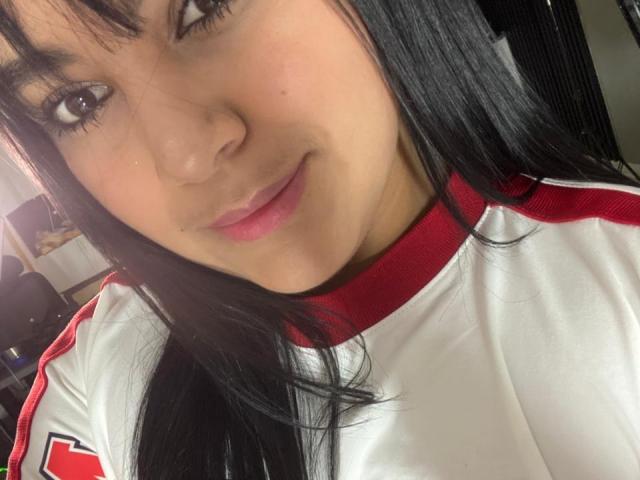 Auroratziu with black hair, pink lips, and light skin, wearing a white and red sports shirt, smiling at the camera. Boobs partially visible. Soft lighting.