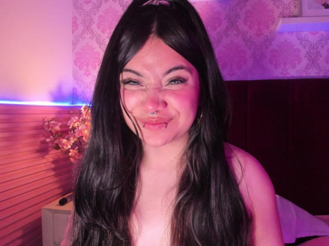 AmaraKazumi mit langen schwarzen Haaren, blauen Augen, Nasenring und Lippenring, lächelnd, die Brüste sichtbar, in einem pink- und violett beleuchteten Schlafzimmer mit floraler Dekoration..