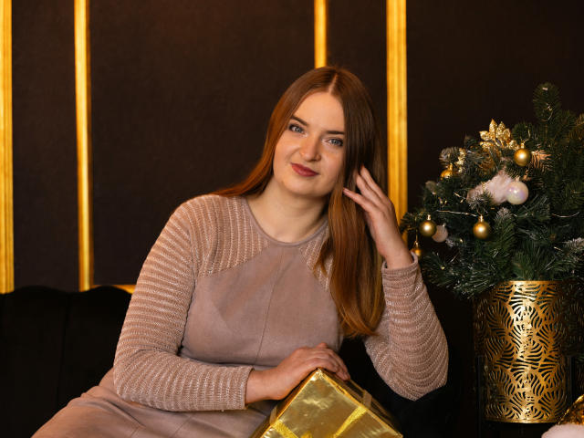 FreyaDak with long red hair, wearing a beige, striped knit dress, holding a gold gift, smiling, right hand touching hair, Christmas tree with gold ornaments on right.