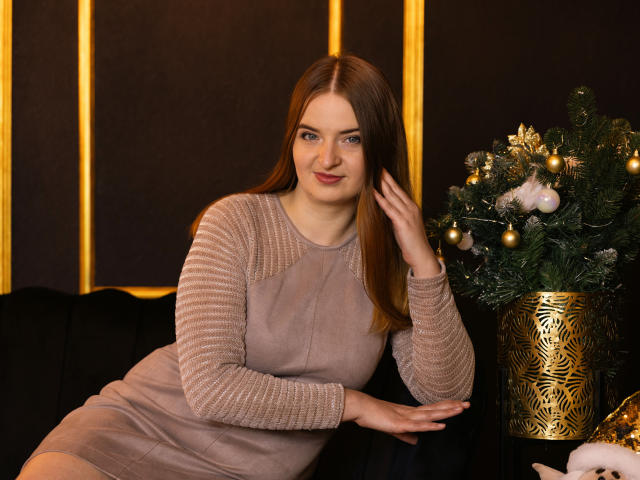 FreyaDak, with brown hair and blue eyes, sits on a black couch in a beige dress, touching her hair, beside a decorated Christmas tree in a gold-patterned pot.